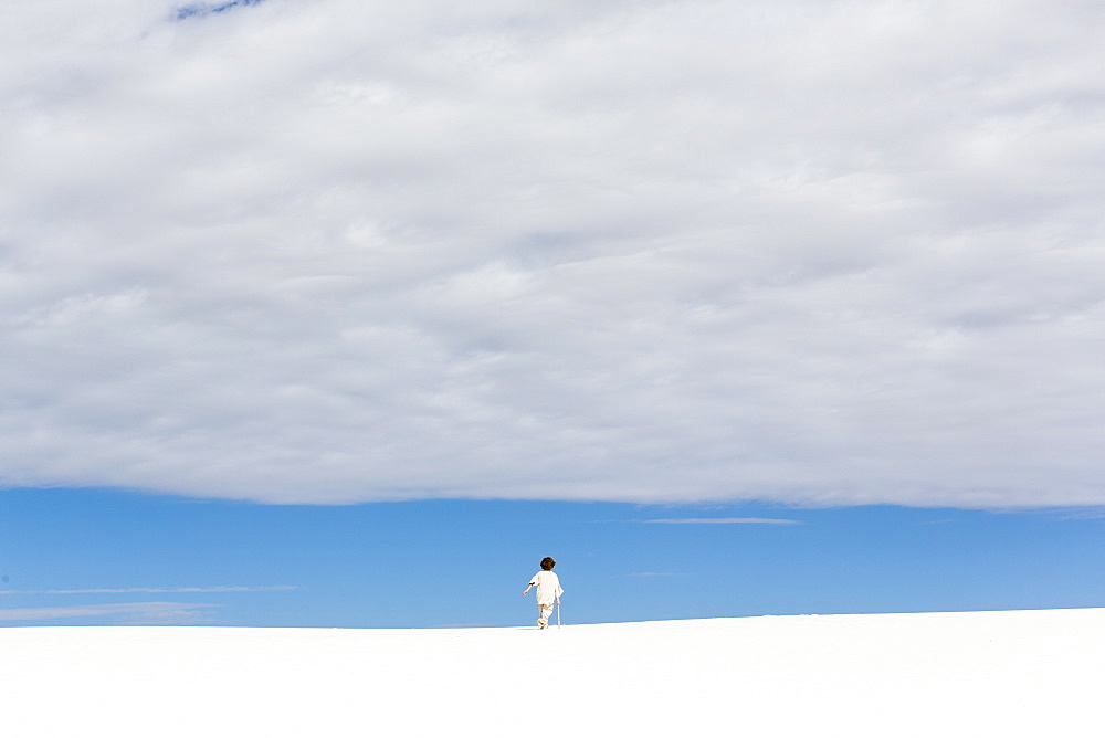 A young boy walking in white sand dunes, a strip of blue sky on the horizon and thick cloud, White Sands National Monument, New Mexico, United States