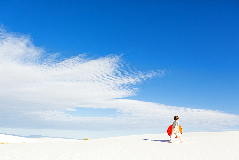 A young boy carrying an orange sled in a white undulating dune landscape, White Sands National Monument, New Mexico, United States