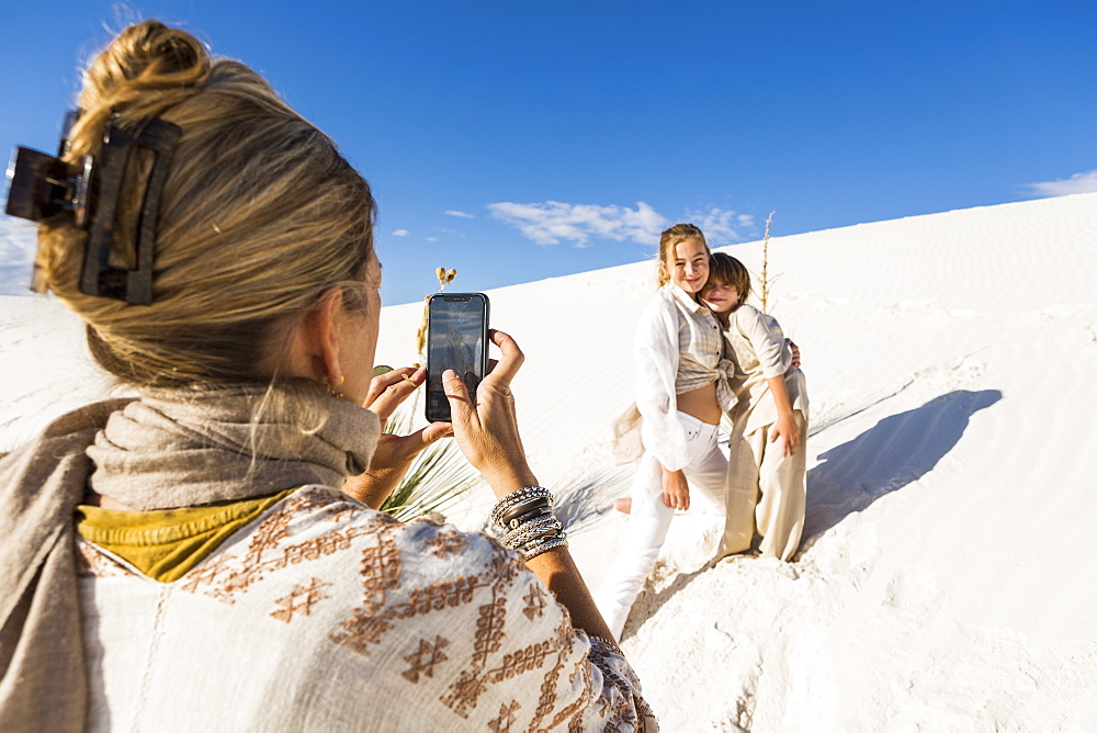 A woman taking picture of her children with a smart phone in white sand dunes landscape under blue sky, White Sands National Monument, New Mexico, United States