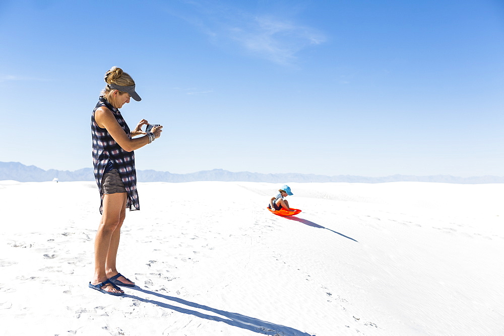 Mother taking picture of son sledding on sand dunes, White Sands National Monument, New Mexico, United States