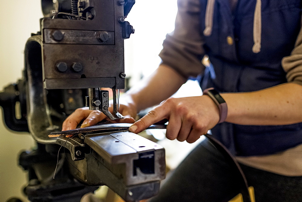 Female saddler standing in workshop, sewing leather strap on saddlery sewing machine, Berkshire, United Kingdom