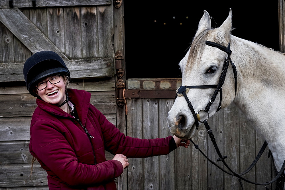 Young woman standing outside stable, holding white Cob horse by it's reins, Berkshire, United Kingdom