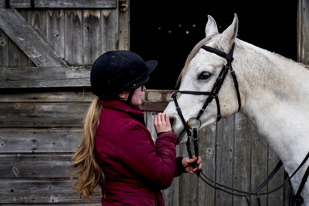 Young woman standing outside stable, holding white Cob horse by it's reins, Berkshire, United Kingdom