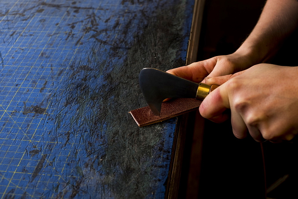 High angle close up of person working in saddler's workshop, using hand tool on leather strap, Berkshire, United Kingdom