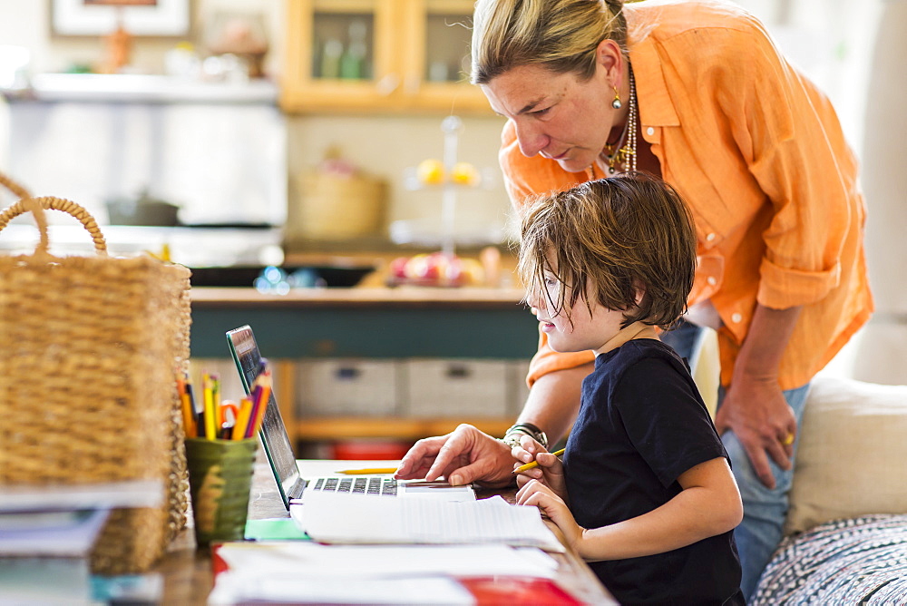 Adult woman helping her six year old son with a remote learning session on a laptop, using a touchpad