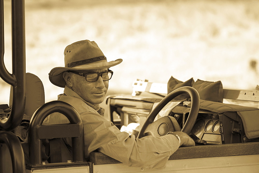 A mature man in hat and glasses in the driving seat of a jeep, sepia tone, Moremi Game Reserve, Botswana