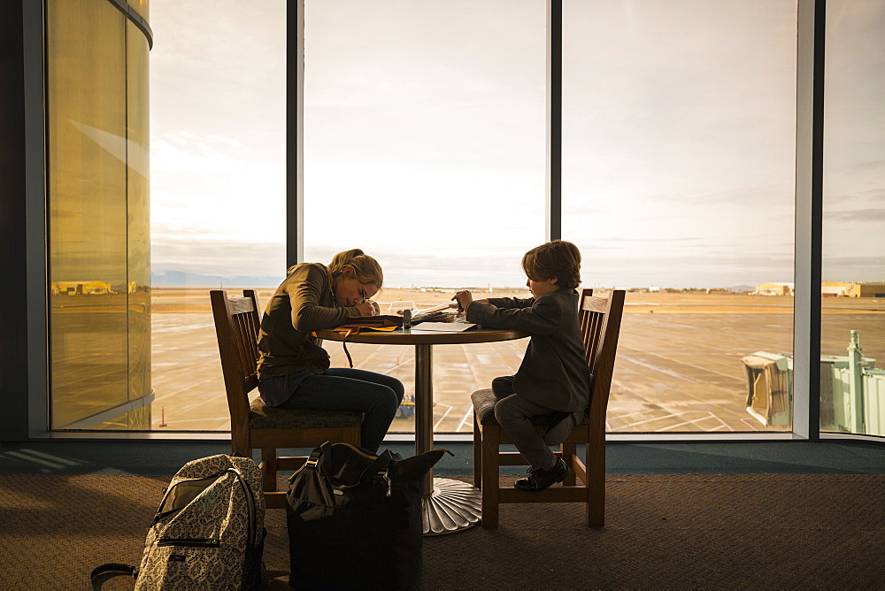 A boy and his older sister seated at a table in an airport lounge, writing and drawing