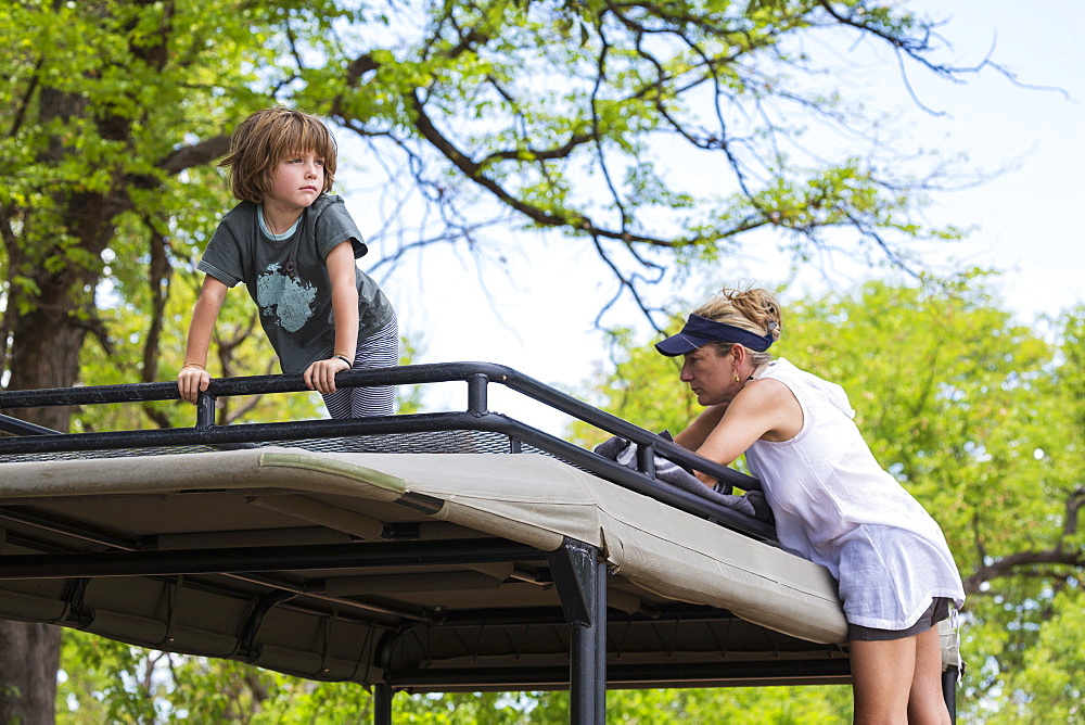 A five year old boy and a teenage girl on the observation platform of a safari jeep under trees, Okavango Delta, Botswana