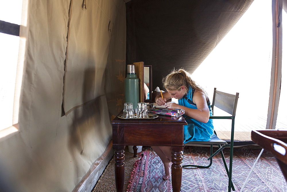 A twelve year old girl seated at a desk in a tent at a wildlife reserve camp, drawing, Okavango Delta, Botswana