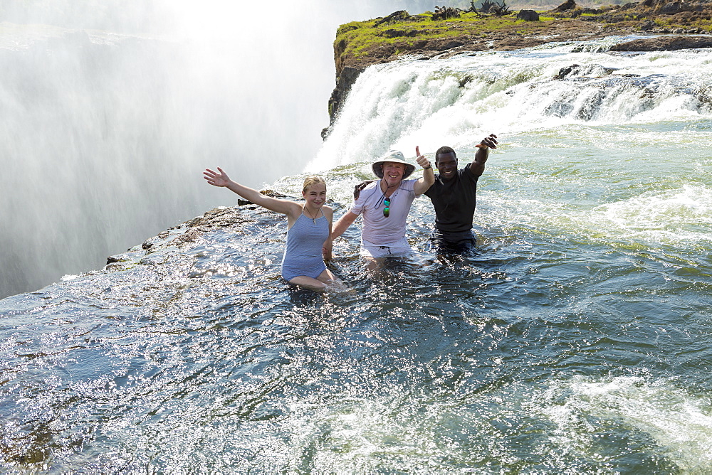 Three people, two men and young teenager standing in the waters of the Devil's Pool on the edge of the Victoria Falls, arms outstretched, Zambia