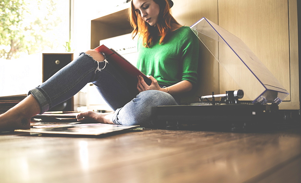 A woman sitting on the floor next to a record player looking at a record sleeve