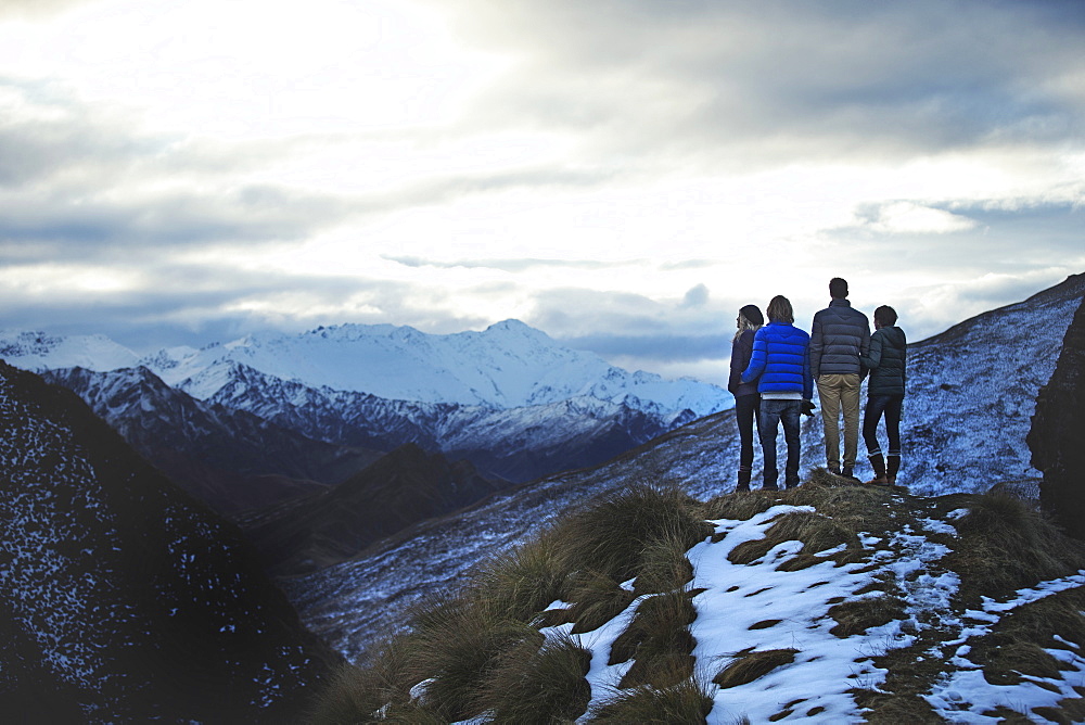 Rear view of four people standing side by side on a mountain, snow-capped peaks in the distance