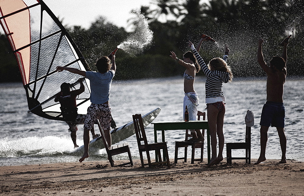 Small group of men and women standing on a sandy beach around table and chairs, arms raised, holding bottles, watching a windsurfer