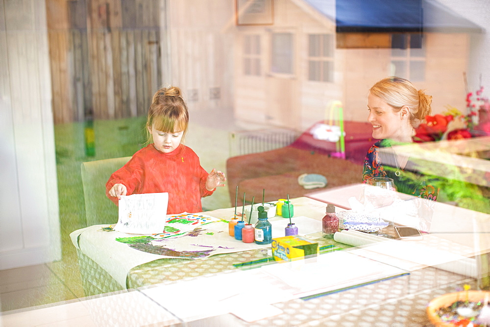 Young girl using paints at kitchen table with mother sitting nearby holding baby