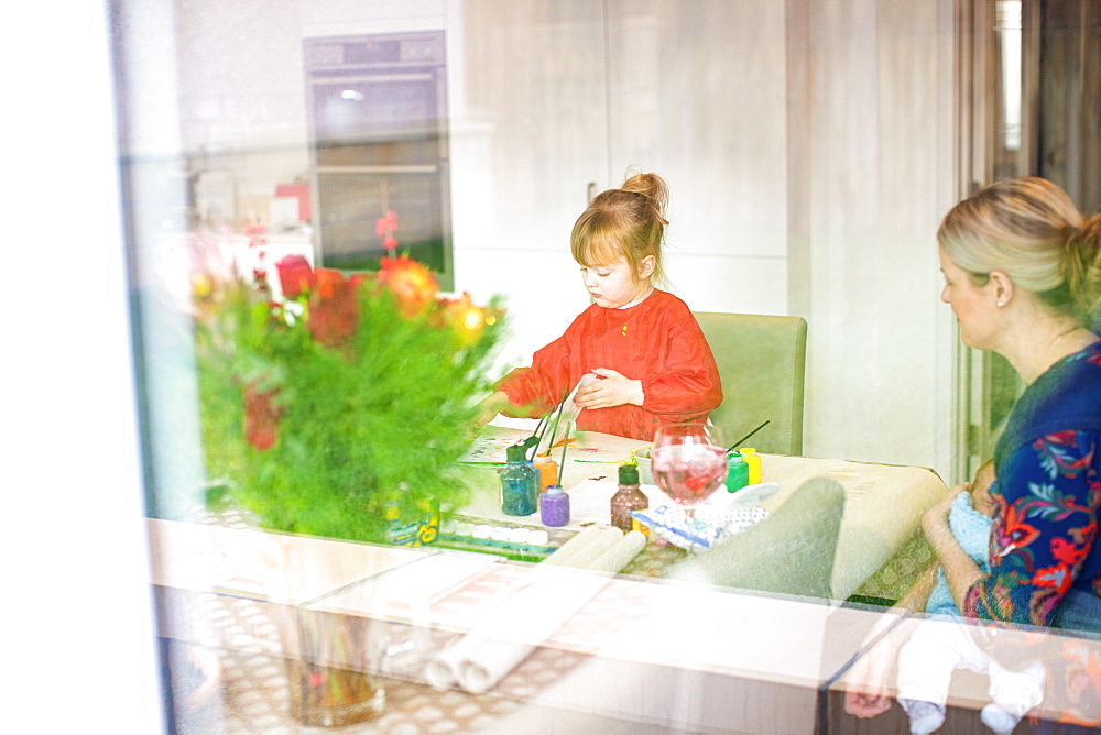 Young girl using paints at kitchen table with mother sitting nearby holding baby