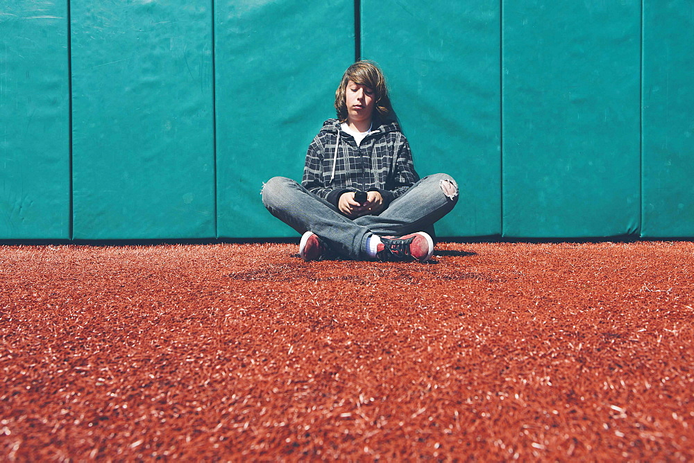 Teenage boy sitting against padded wall at sports field holding mobile phone