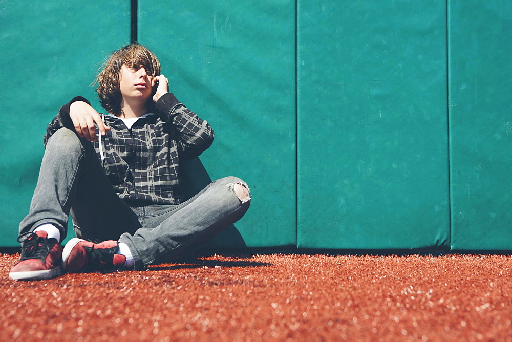 Teenage boy sitting against padded wall at sports field holding mobile phone