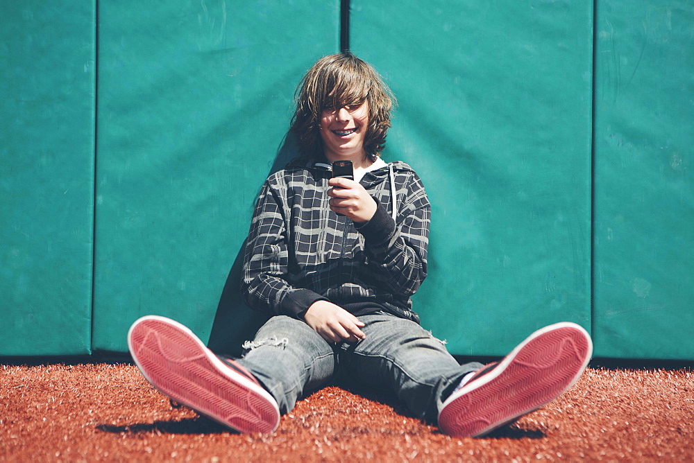 Teenage boy sitting against padded wall at sports field holding mobile phone