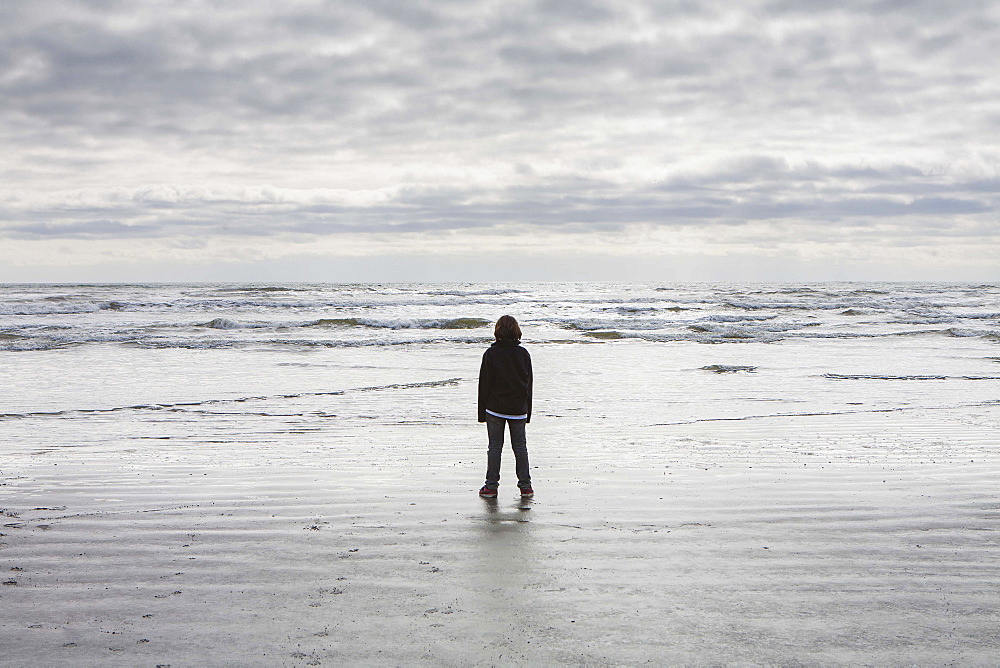 Teenage boy standing on vast beach waves and overcast sky in distance, Olympic National Park, Washington, United States of America