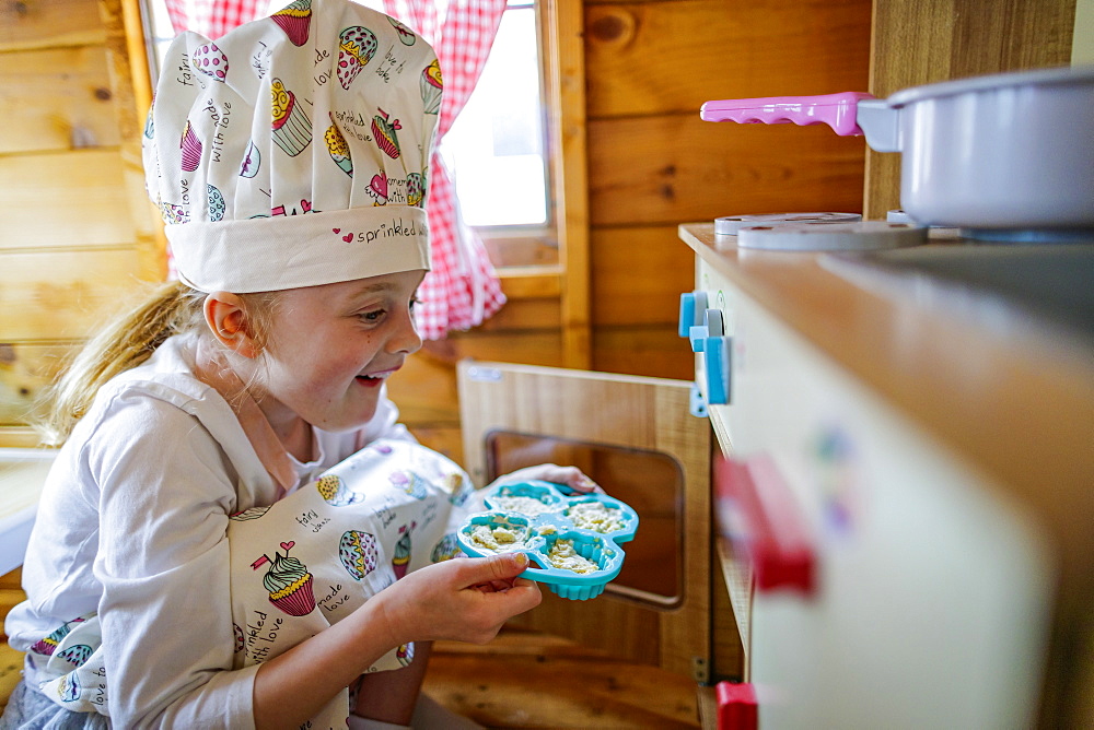 Young girl in wendy house putting cup cakes in oven pretending to cook in kitchen