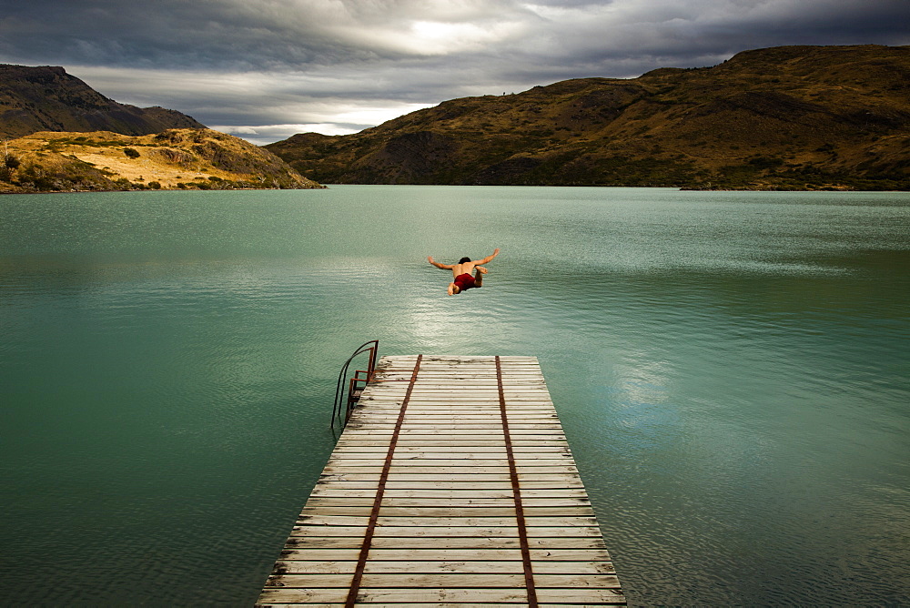 A young man in mid air, diving off a wooden pier, into calm lake surrounded by mountains in Torres del Paine National Park, Chile, Torres del Paine National Park, Chile.