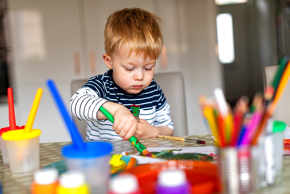 Three year old boy busy painting at home, with paint pots and brushes, Scotland, United Kingdom