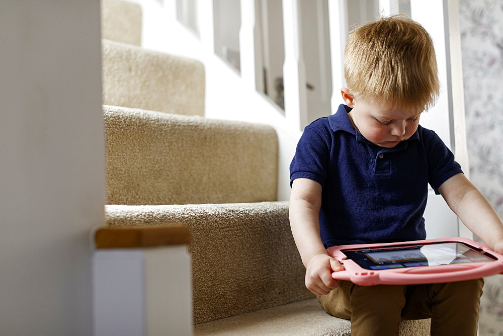 Three year old boy seated on the stairs looking at a digital tablet, Scotland, United Kingdom