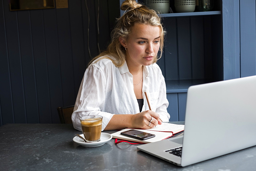 Woman sitting alone at a cafe table with a laptop computer, writing in note book, working remotely, Watlington, Oxfordshire, United Kingdom