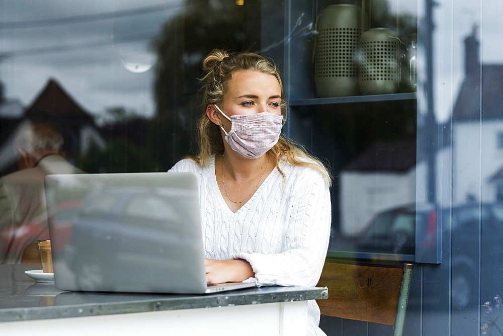 Woman wearing face mask sitting alone at a cafe table with a laptop computer, working remotely, Watlington, Oxfordshire, United Kingdom