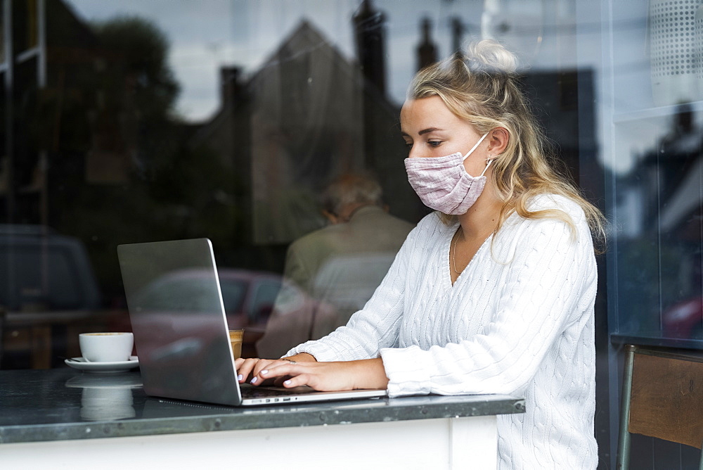 Woman wearing face mask sitting alone at a cafe table with a laptop computer, working remotely, Watlington, Oxfordshire, United Kingdom