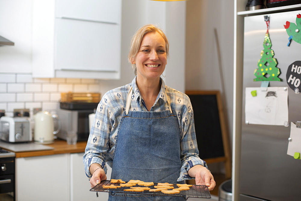 Blond woman standing in kitchen, holding tray with Christmas cookies, smiling at camera