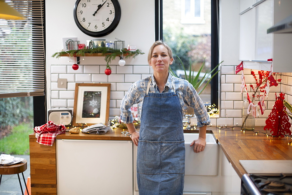 Blond woman wearing blue apron standing in kitchen, smiling at camera