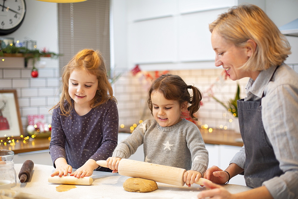 Blond woman wearing blue apron and two girls standing in kitchen, baking Christmas cookies