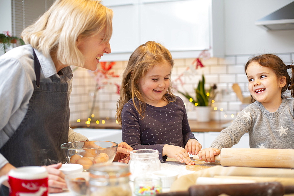 Blond woman wearing blue apron and two girls standing in kitchen, baking Christmas cookies