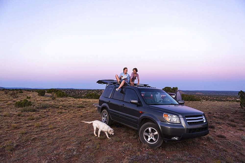 Teenage girl and her younger brother sitting atop their SUV at sundown