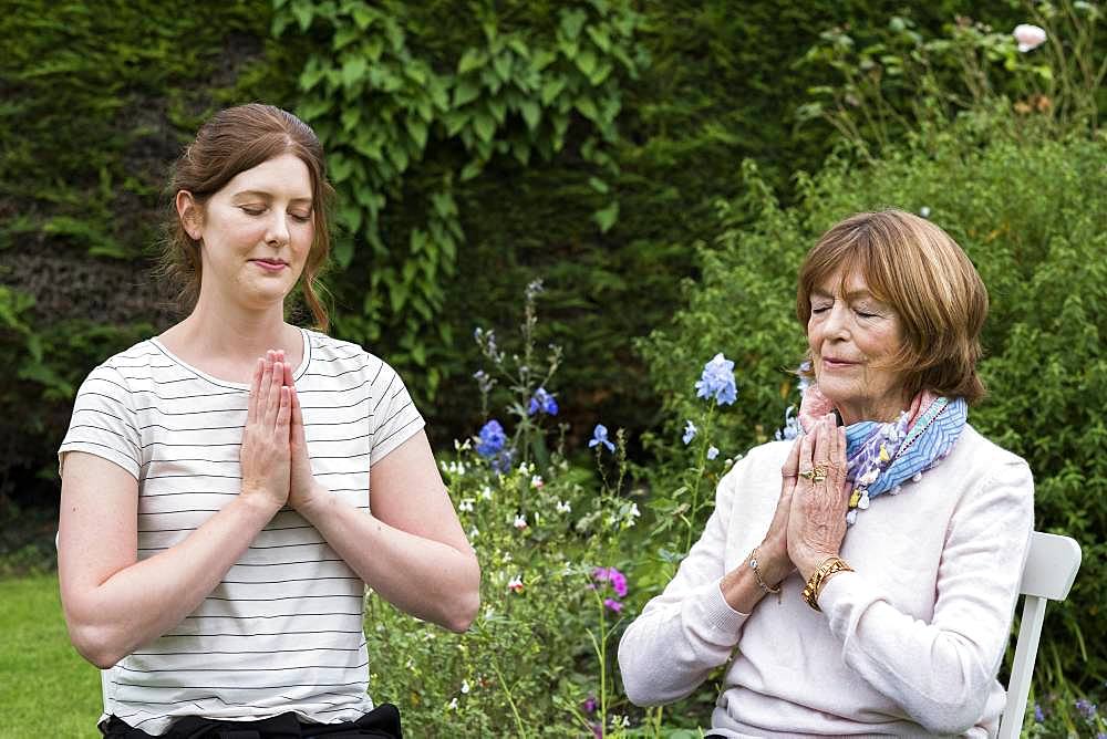 Woman and female therapist seated in a garden, hands together and eyes closed