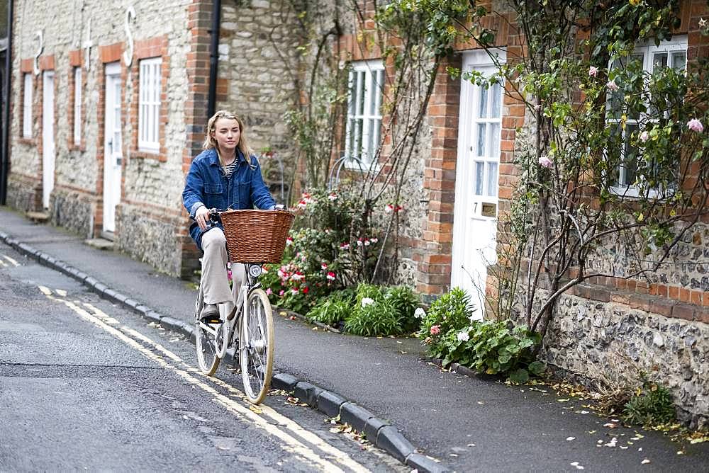 Young blond woman cycling down a village street, Oxfordshire, England, United Kingdom