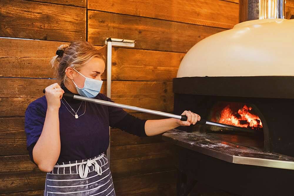 Waitress wearing blue striped apron and face mask standing at a pizza oven in a restaurant