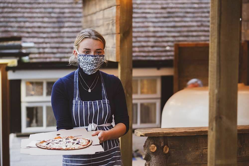 Woman waitress in apron and face mask holding plate of pizza