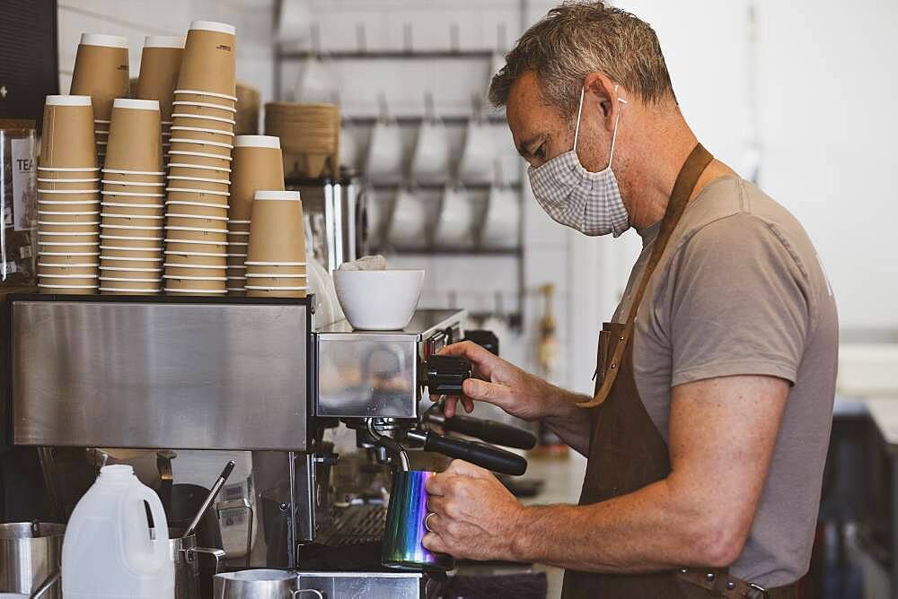 Male barista wearing brown apron and face mask working in a cafe, making espresso