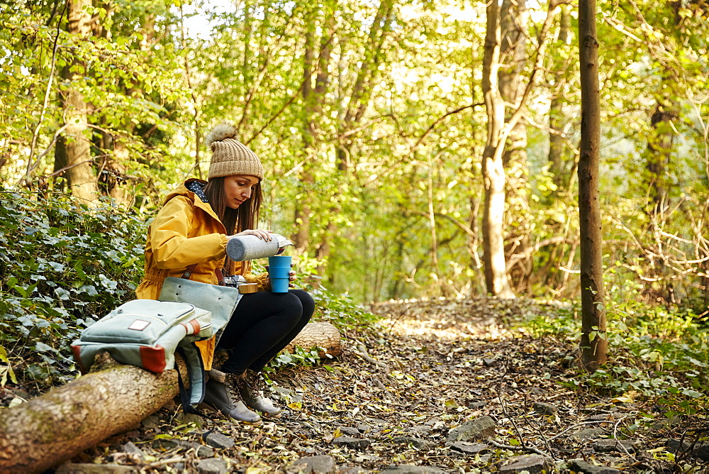 Woman sitting on tree trunk in woodland pouring drink from thermos flask