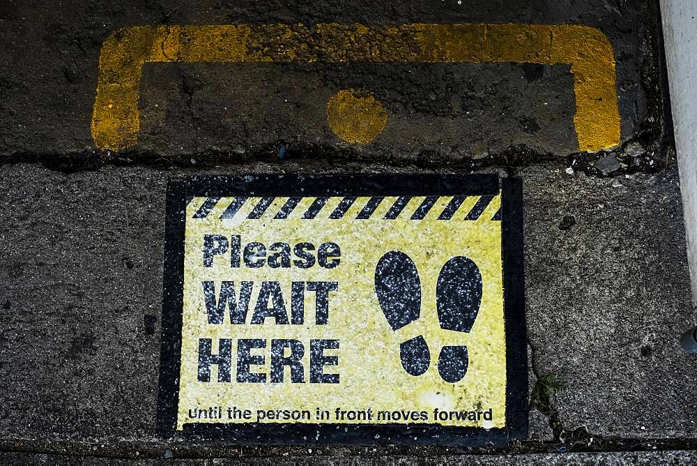 High angle close up of black and yellow 'Please Wait Here' sign on asphalt ground.