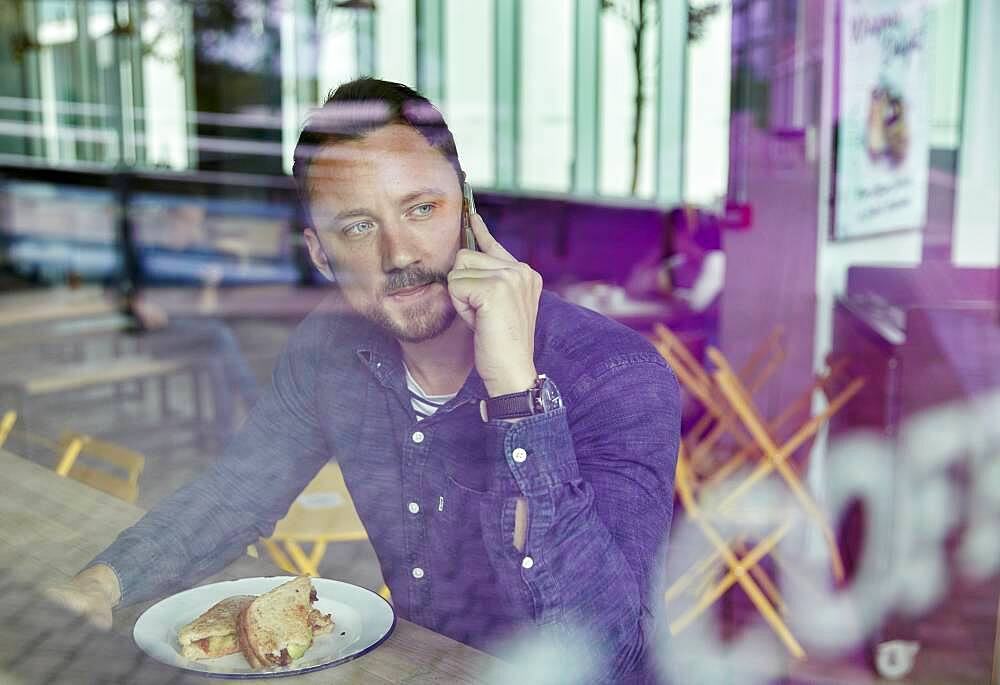 Man in a face mask seated at a cafe table using a mobile phone, view through a window
