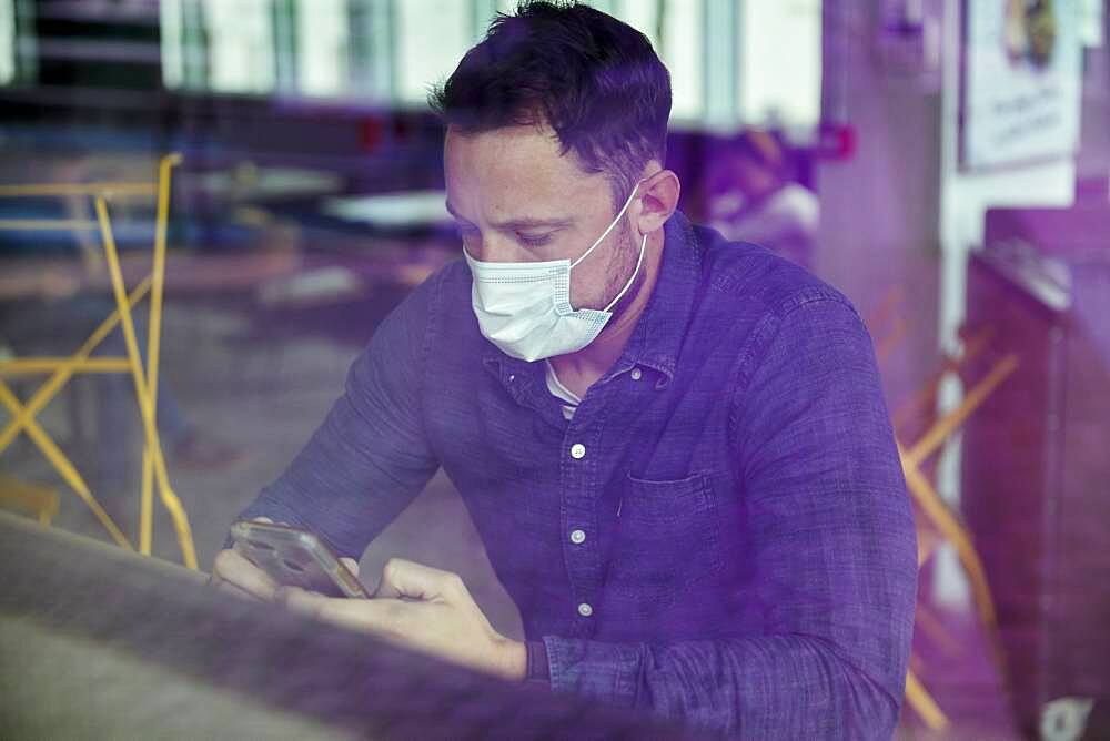 Man in a face mask seated at a cafe table using a mobile phone, view through a window