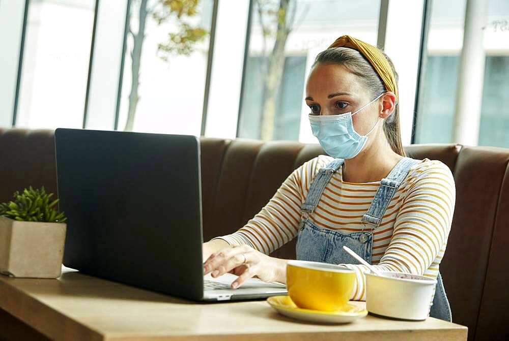 Woman wearing a face mask seated in a cafe using a laptop