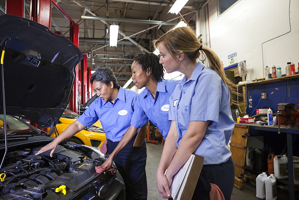 Three female mechanics look inside the engine compartment of a car in a repair shop