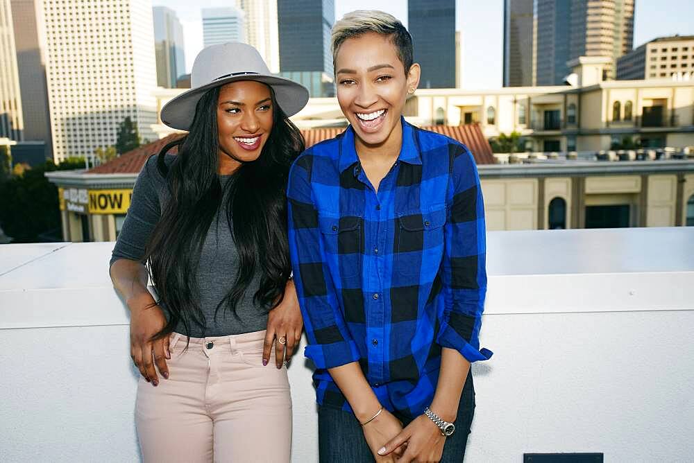 Two young women on a rooftop posing for photographs, city skyline background.