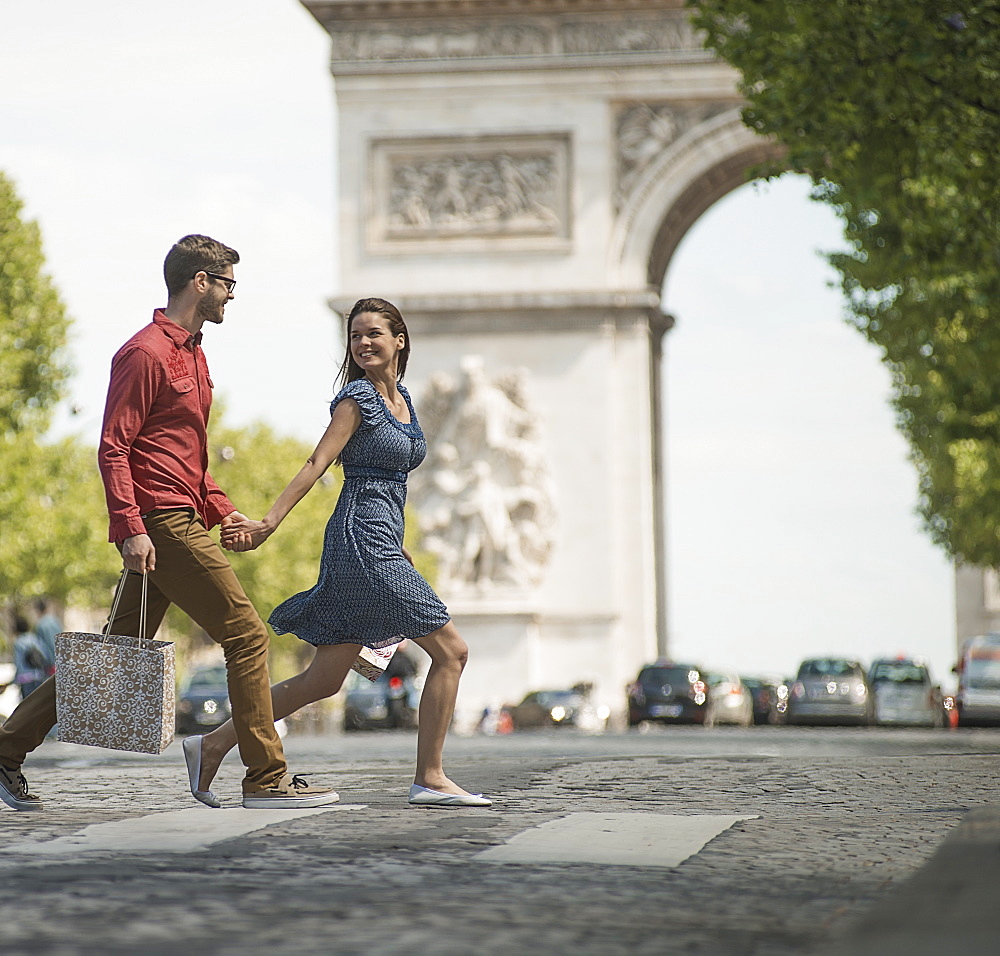 A couple hand in hand carrying shopping bags and crossing the road by a historic monument in the heart of a European city, France