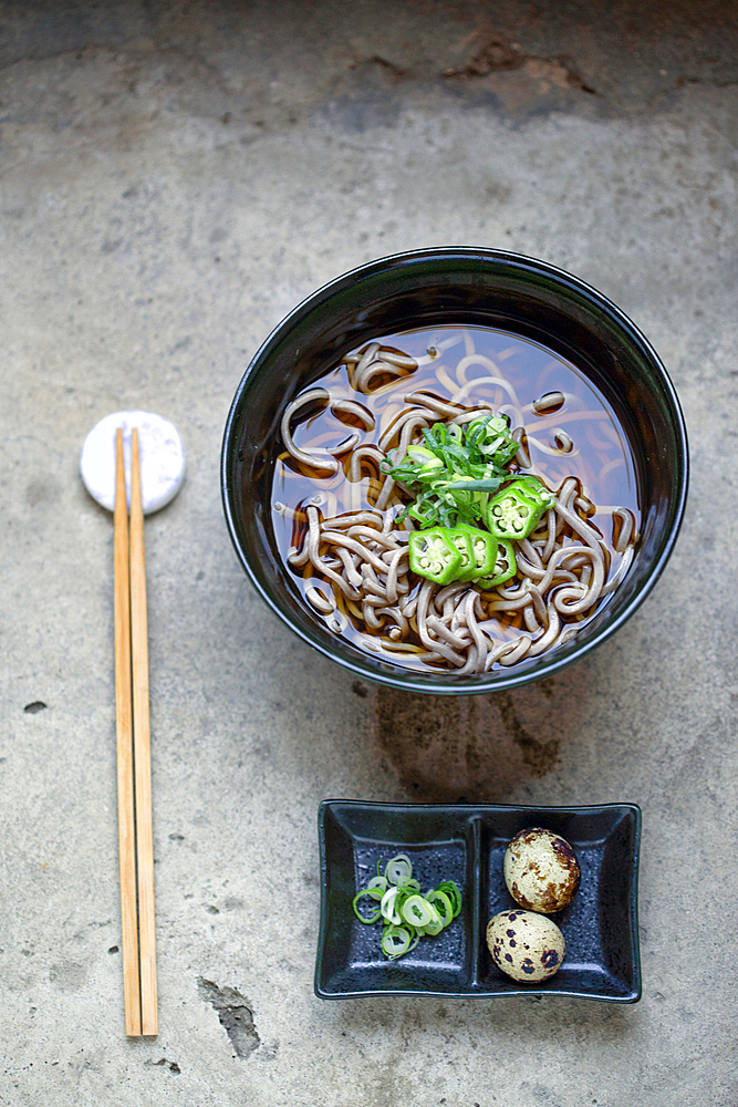 A black china bowl of noodles and broth, sliced green chilli and a dish with quail's eggs and sliced vegetables.