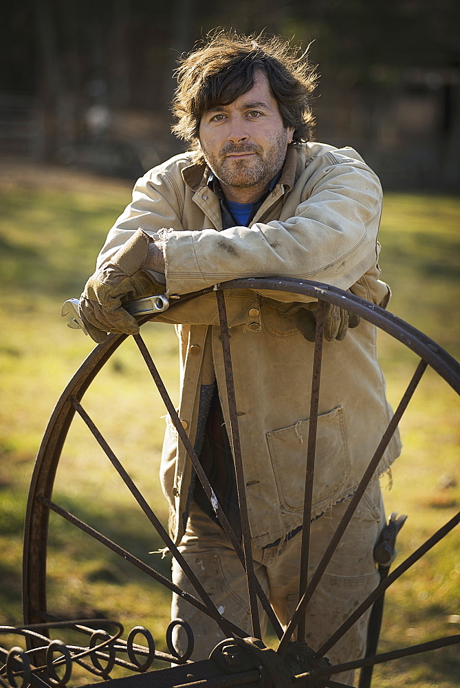 Working on an organic farm. A man in overalls wearing work gloves, leaning on a round metal wheel with spokes, Kerhonkson, New York, USA